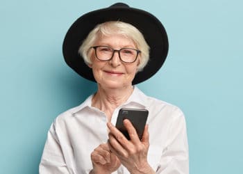 Senior positive wrinkled woman pleased with new useful features of modern gadget, holds cellular with touchscreen, wears stylish black hat, elegant white shirt black trousers with red belt. technology
