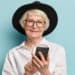 Senior positive wrinkled woman pleased with new useful features of modern gadget, holds cellular with touchscreen, wears stylish black hat, elegant white shirt black trousers with red belt. technology